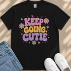 A black t-shirt with a colorful graphic design that says "KEEP GOING CUTIE" surrounded by flowers and hearts. The t-shirt is placed next to a pair of jeans and white sneakers.
