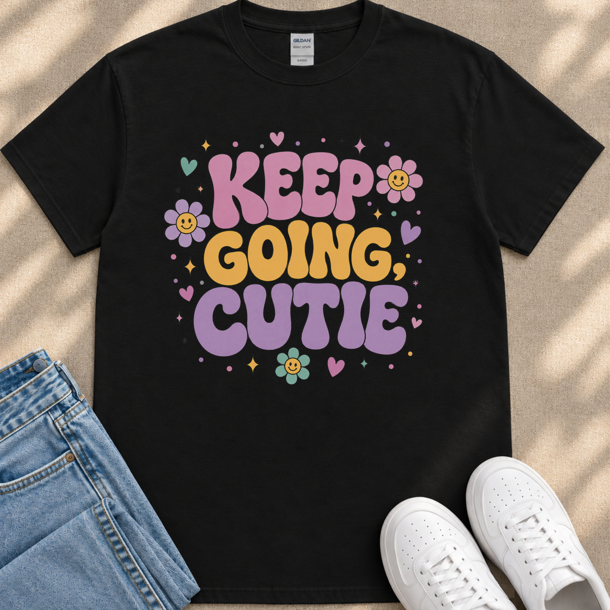 A black t-shirt with a colorful graphic design that says "KEEP GOING CUTIE" surrounded by flowers and hearts. The t-shirt is placed next to a pair of jeans and white sneakers.