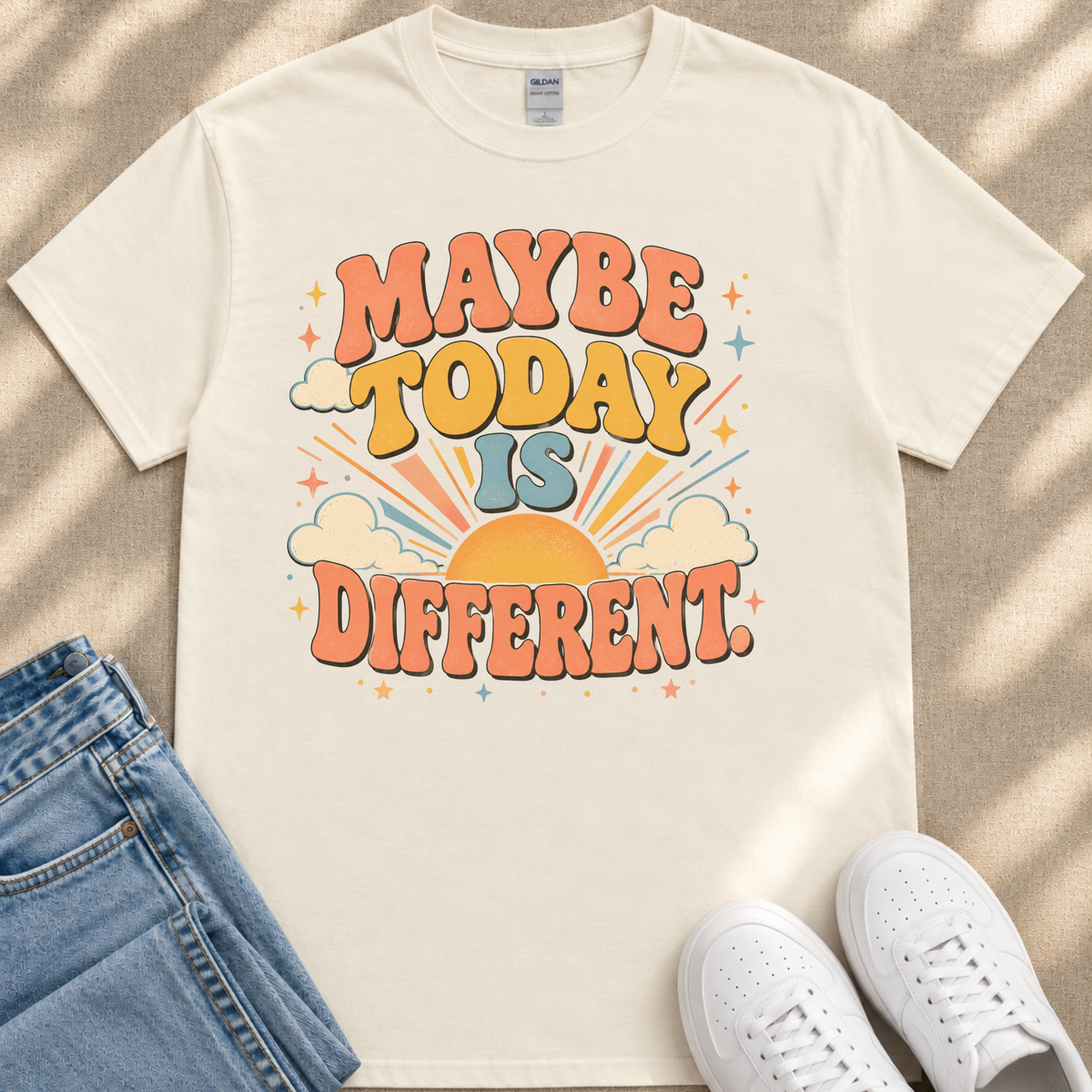 A white t-shirt with a colorful graphic design that says "MAYBE TODAY IS DIFFERENT" along with a sun and clouds design. The t-shirt is placed on a beige surface, and there are a pair of blue jeans and white sneakers visible in the image.