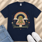 A navy blue t-shirt with a whimsical graphic design depicting a frog sitting on a mushroom, holding a cup, and surrounded by a rainbow, flowers, and a sun. The text on the t-shirt reads "Small steps still count".
