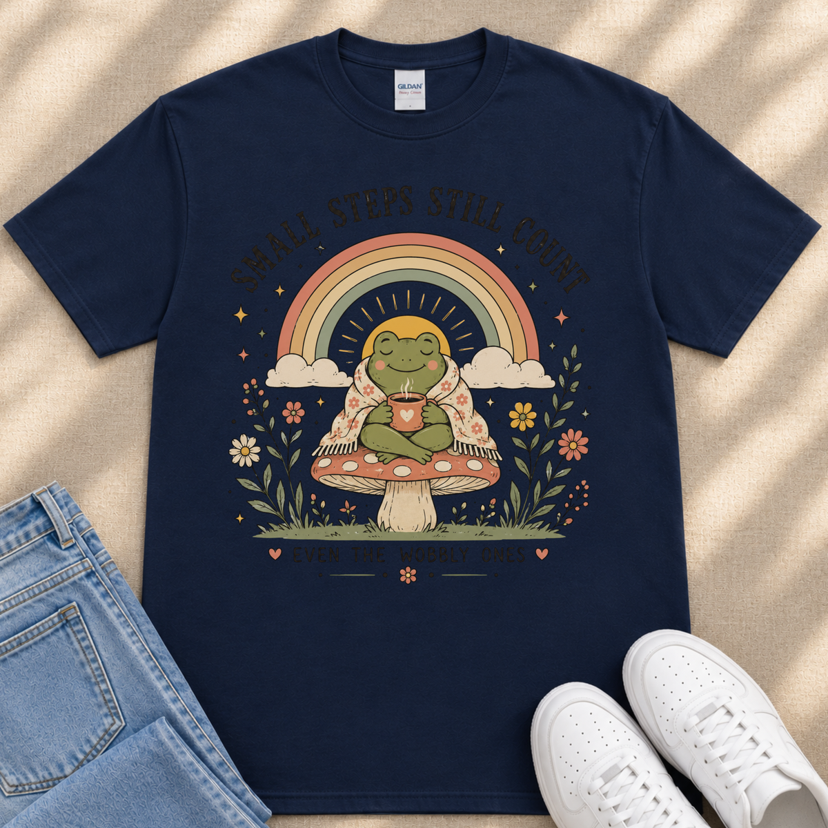 A navy blue t-shirt with a whimsical graphic design depicting a frog sitting on a mushroom, holding a cup, and surrounded by a rainbow, flowers, and a sun. The text on the t-shirt reads "Small steps still count".