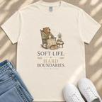 A white t-shirt with a graphic design depicting a bear sitting in a rocking chair, holding a cup and surrounded by potted plants. The text on the t-shirt reads "Soft Life, Hard Boundaries."