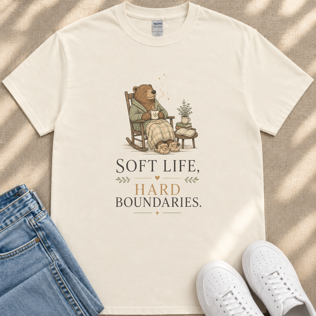 A white t-shirt with a graphic design depicting a bear sitting in a rocking chair, holding a cup and surrounded by potted plants. The text on the t-shirt reads "Soft Life, Hard Boundaries."