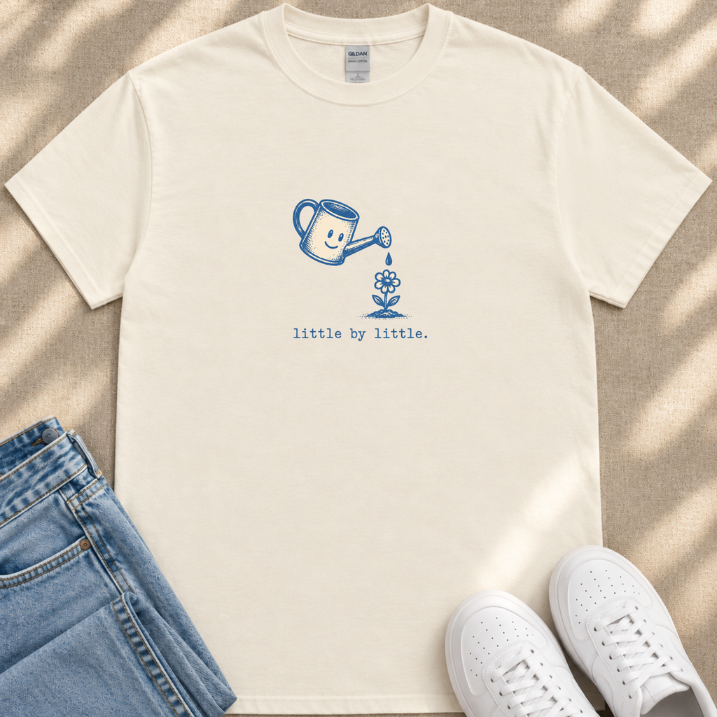 A white t-shirt with a blue watering can graphic and the text "little by little" printed on it, along with a pair of blue jeans and white sneakers.