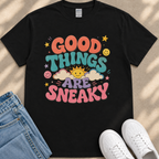 A black t-shirt with a colorful graphic design that says "GOOD THINGS ARE SNAKY" along with various cheerful elements such as smiley faces, clouds, and stars.