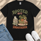 a black t-shirt with a graphic design depicting a potted plant, a stack of books, and a pair of headphones, along with the text "ROOTED BUT UNBOTHERED" printed on it.