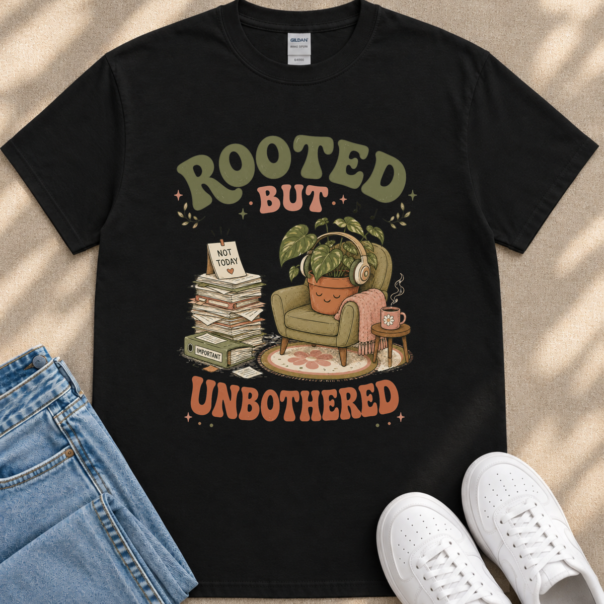 a black t-shirt with a graphic design depicting a potted plant, a stack of books, and a pair of headphones, along with the text "ROOTED BUT UNBOTHERED" printed on it.