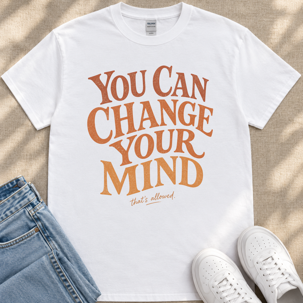 A white t-shirt with the text "You Can Change Your Mind" printed on it, along with the phrase "that's allowed." The t-shirt is placed on a pair of jeans and white sneakers, suggesting a casual outfit.