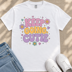 A white t-shirt with a colorful graphic design featuring the text "KEEP GOING CUTIE" surrounded by flowers, hearts, and stars.