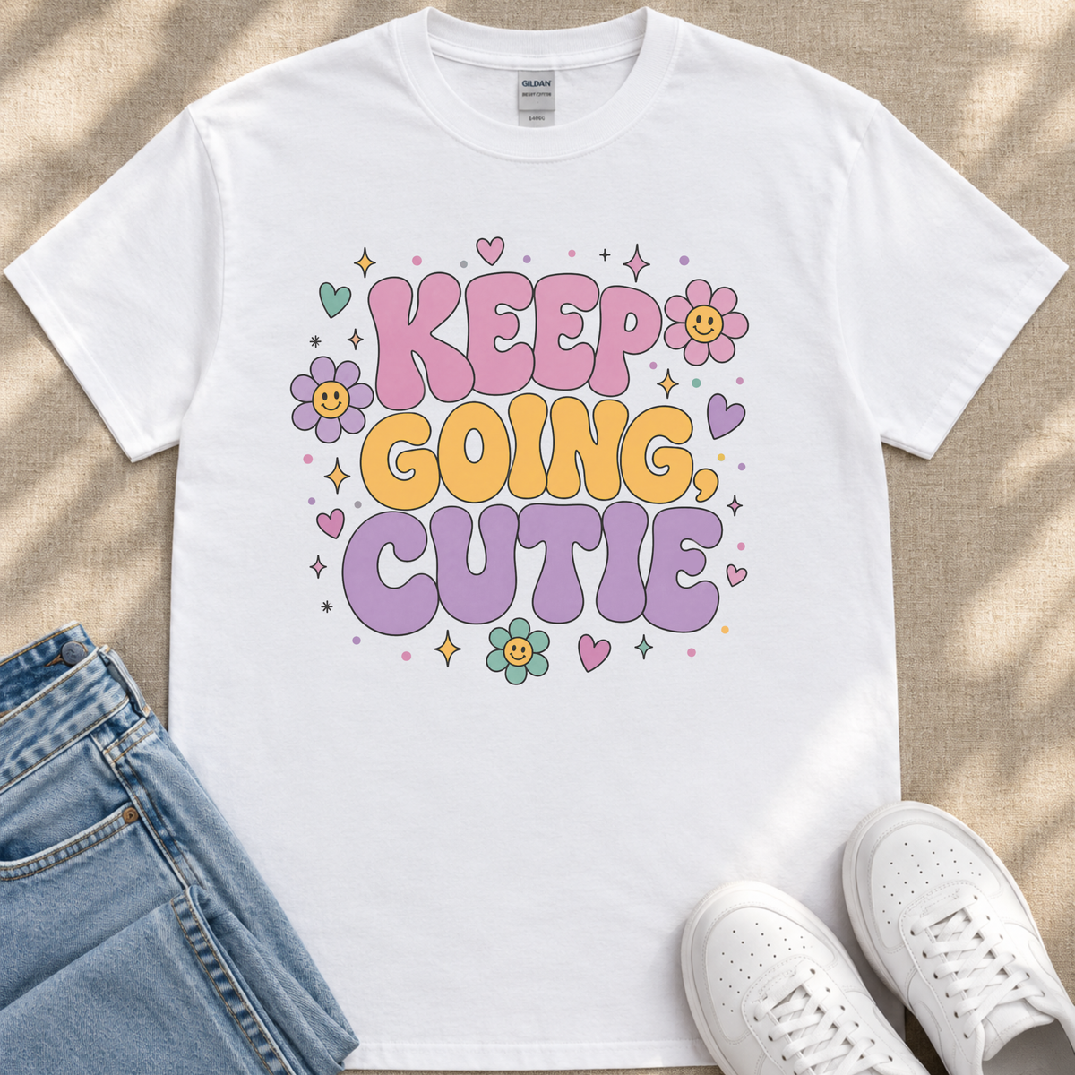 A white t-shirt with a colorful graphic design featuring the text "KEEP GOING CUTIE" surrounded by flowers, hearts, and stars.