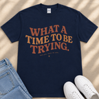 A navy blue t-shirt with the text "What a TIME to BE TRYING." printed on it, along with the phrase "and get, here we are." placed on a beige surface next to a pair of blue jeans and white sneakers.
