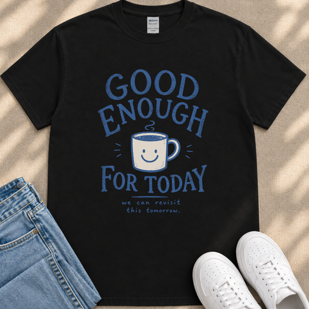 A black t-shirt with a blue graphic design that says "GOOD ENOUGH FOR TODAY" and features a smiling cup. The t-shirt is placed on a beige surface, and there are a pair of blue jeans and white sneakers visible in the image.