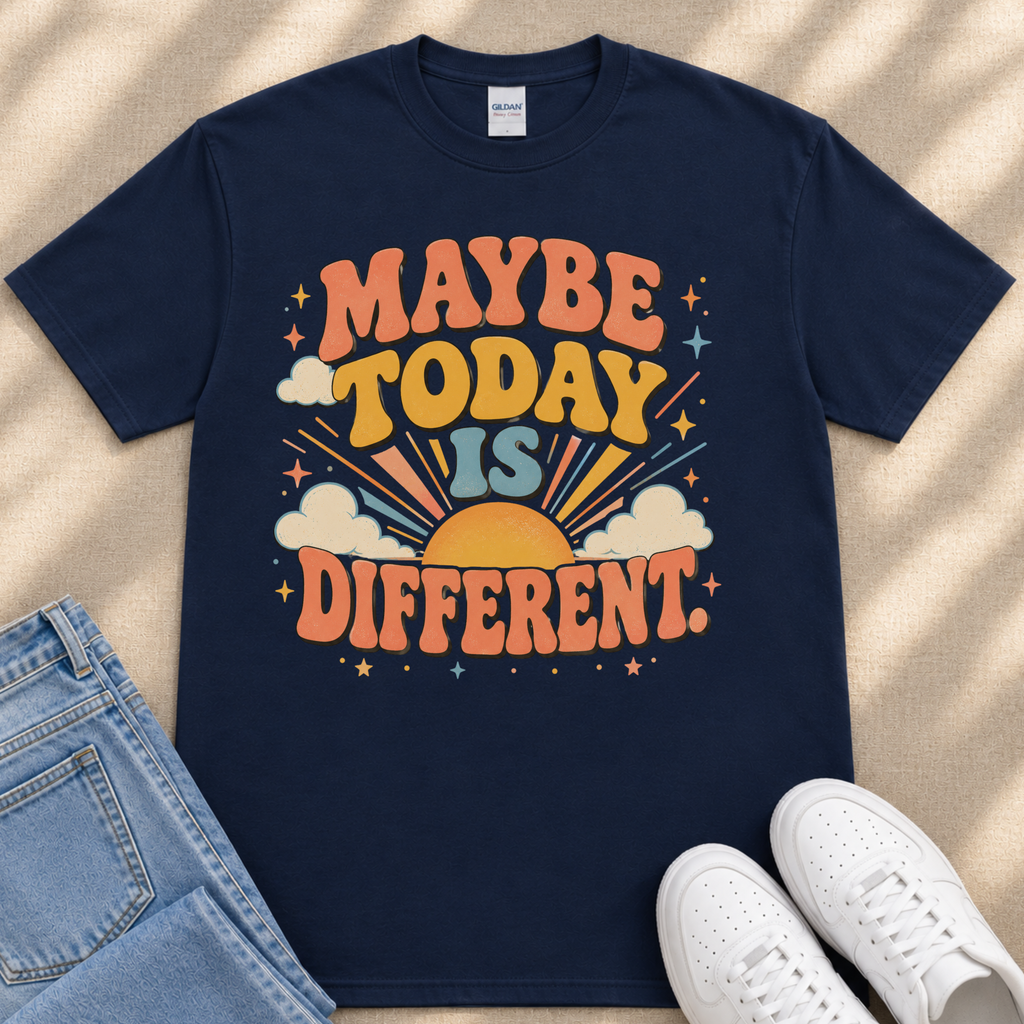A navy blue t-shirt with a colorful graphic design that says "MAYBE TODAY IS DIFFERENT" and features a sun, clouds, and stars. The t-shirt is placed on a beige surface, and there are a pair of jeans and white sneakers visible in the image.