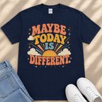A navy blue t-shirt with a colorful graphic design that says "MAYBE TODAY IS DIFFERENT" and features a sun, clouds, and stars. The t-shirt is placed on a beige surface, and there are a pair of jeans and white sneakers visible in the image.