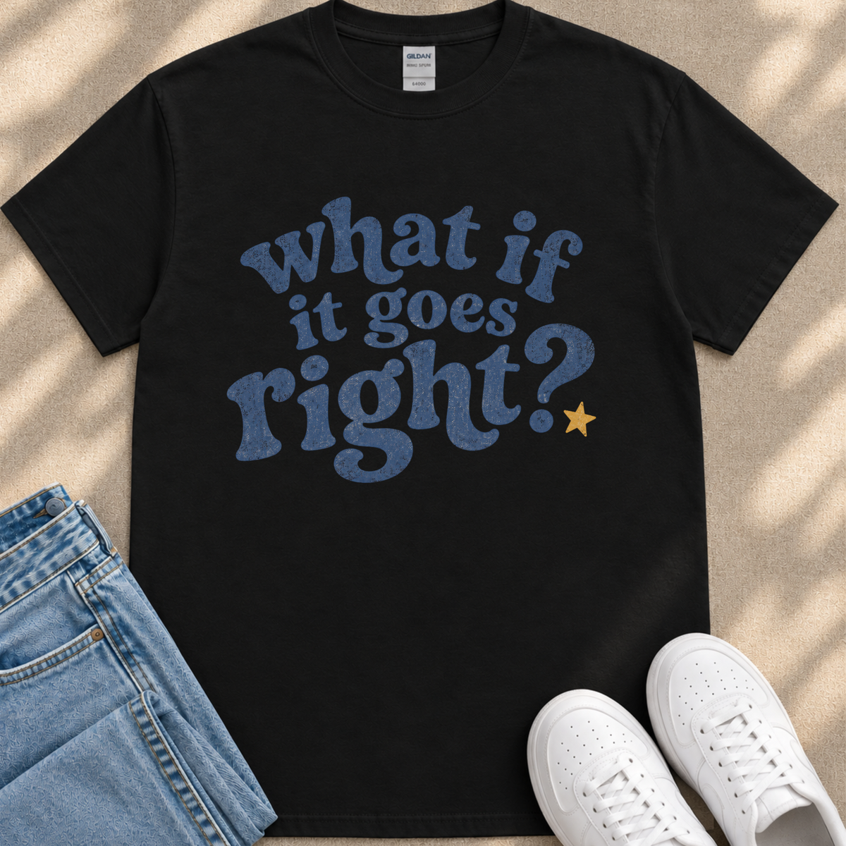 A black t-shirt with a blue text design that reads "What if it goes right?" along with a yellow star, placed on a beige surface next to a pair of blue jeans and white sneakers.