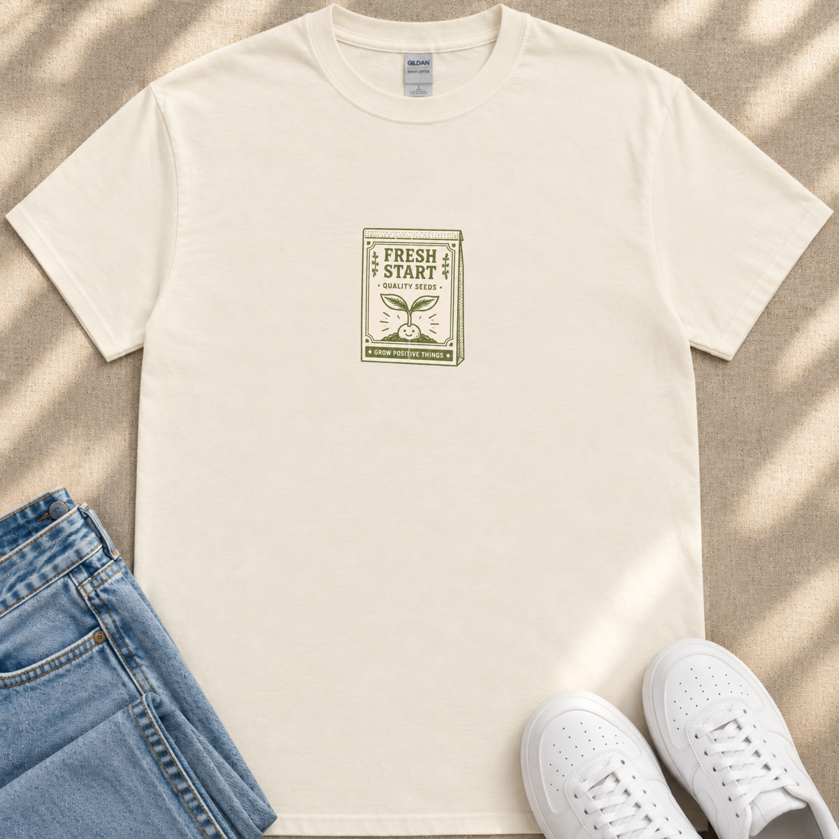 A white t-shirt with a graphic design, a pair of blue jeans, and white sneakers are arranged on a beige surface.