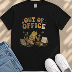 A black t-shirt with a colorful graphic design depicting a frog sitting on a laptop, surrounded by flowers and butterflies, with the text "Out of Office" written above.