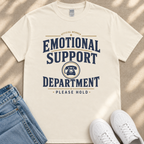 a white t-shirt with a graphic design that reads "Emotional Support Department Please Hold" in blue and yellow text. The t-shirt is placed on a beige surface, and there are a pair of blue jeans and white sneakers visible in the background.