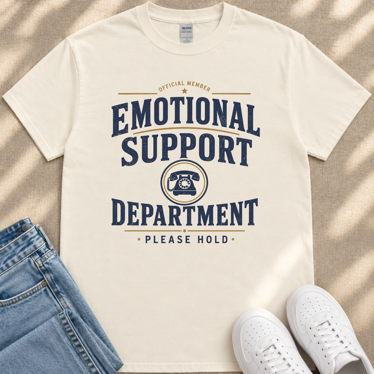 a white t-shirt with a graphic design that reads "Emotional Support Department Please Hold" in blue and yellow text. The t-shirt is placed on a beige surface, and there are a pair of blue jeans and white sneakers visible in the background.
