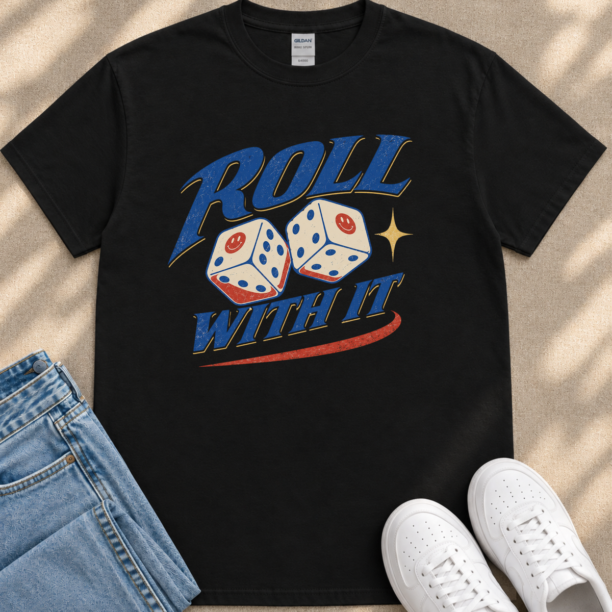 A black t-shirt with a graphic design featuring two dice with a smiley face, along with the text "Roll with it" and a red and blue stripe. The t-shirt is placed next to a pair of jeans and white sneakers.