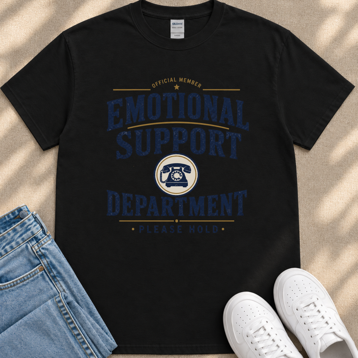 a black t-shirt with a graphic design that reads "Emotional Support Department Please Hold" in blue and yellow text. The t-shirt is placed on a beige surface, and there are a pair of blue jeans and white sneakers visible in the image.