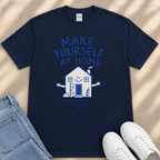 A navy blue t-shirt with a graphic design of a house and the text "MAKE YOURSELF AT HOME" printed on it, along with a smaller text below that says "and you'll happen to be." The t-shirt is laid out on a beige surface, and there are a pair of blue jeans and white sneakers visible in the image.