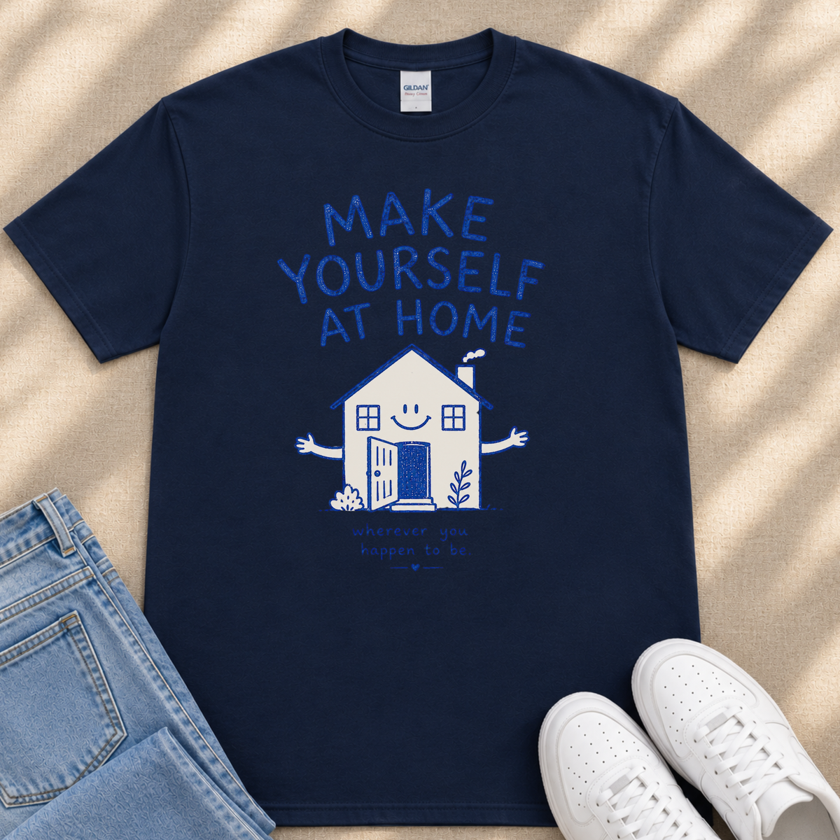 A navy blue t-shirt with a graphic design of a house and the text "MAKE YOURSELF AT HOME" printed on it, along with a smaller text below that says "and you'll happen to be." The t-shirt is laid out on a beige surface, and there are a pair of blue jeans and white sneakers visible in the image.