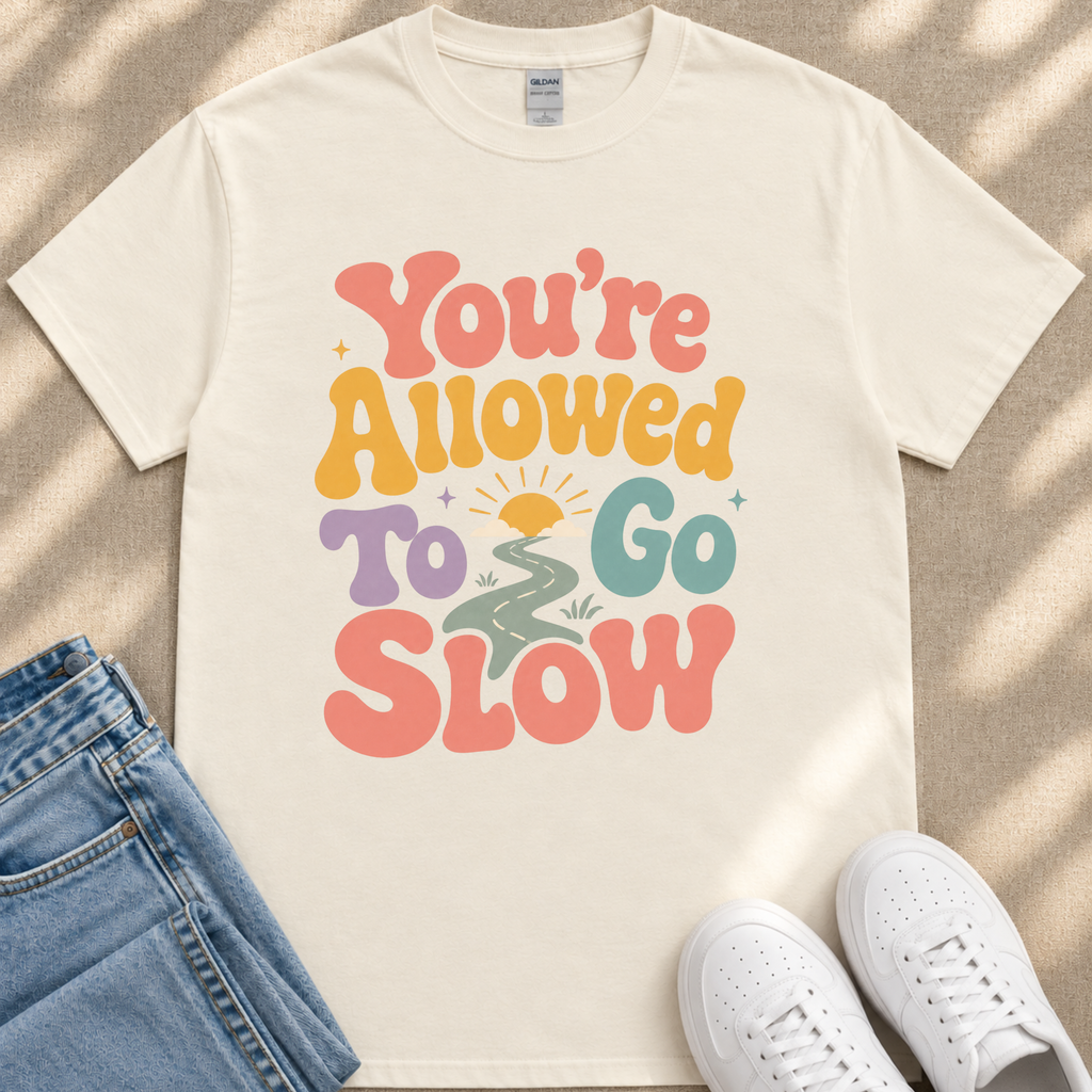 A white t-shirt with a colorful graphic design that says "You're Allowed to Go Slow" printed on it. The t-shirt is placed on a beige surface, and there are a pair of blue jeans and white sneakers visible in the image.