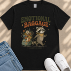 a black t-shirt with a graphic design depicting a raccoon pulling a suitcase filled with various items, including flowers and snacks. The text on the t-shirt reads "Emotional Baggage" and "Carry-on Size".