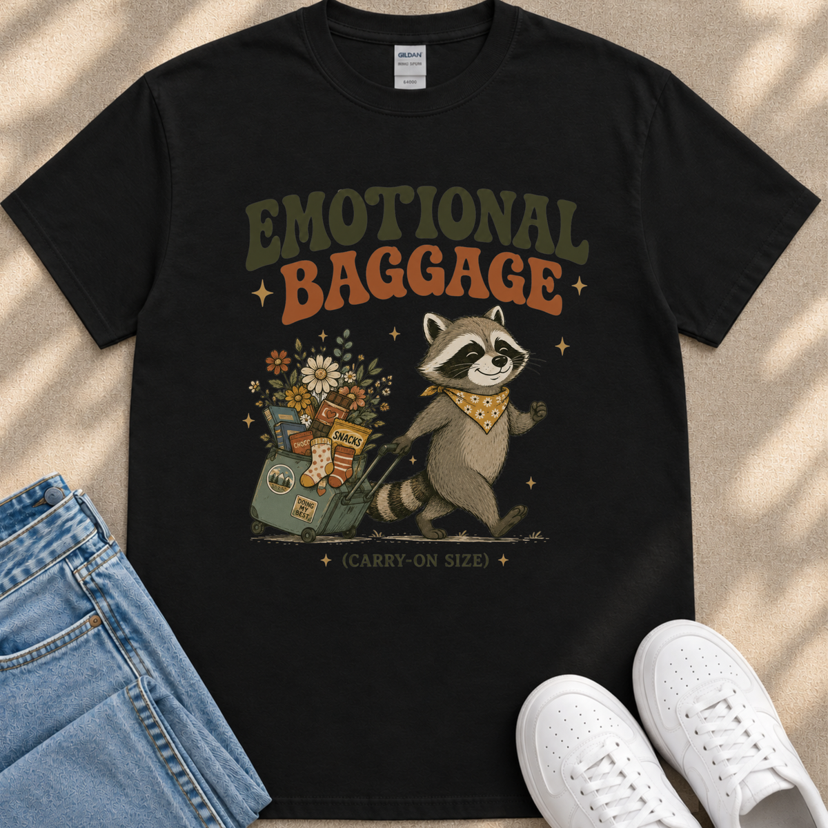 a black t-shirt with a graphic design depicting a raccoon pulling a suitcase filled with various items, including flowers and snacks. The text on the t-shirt reads "Emotional Baggage" and "Carry-on Size".
