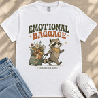 A white t-shirt with a graphic design depicting a raccoon pulling a suitcase filled with various items, accompanied by the text "Emotional Baggage" and "Carry-on Size".