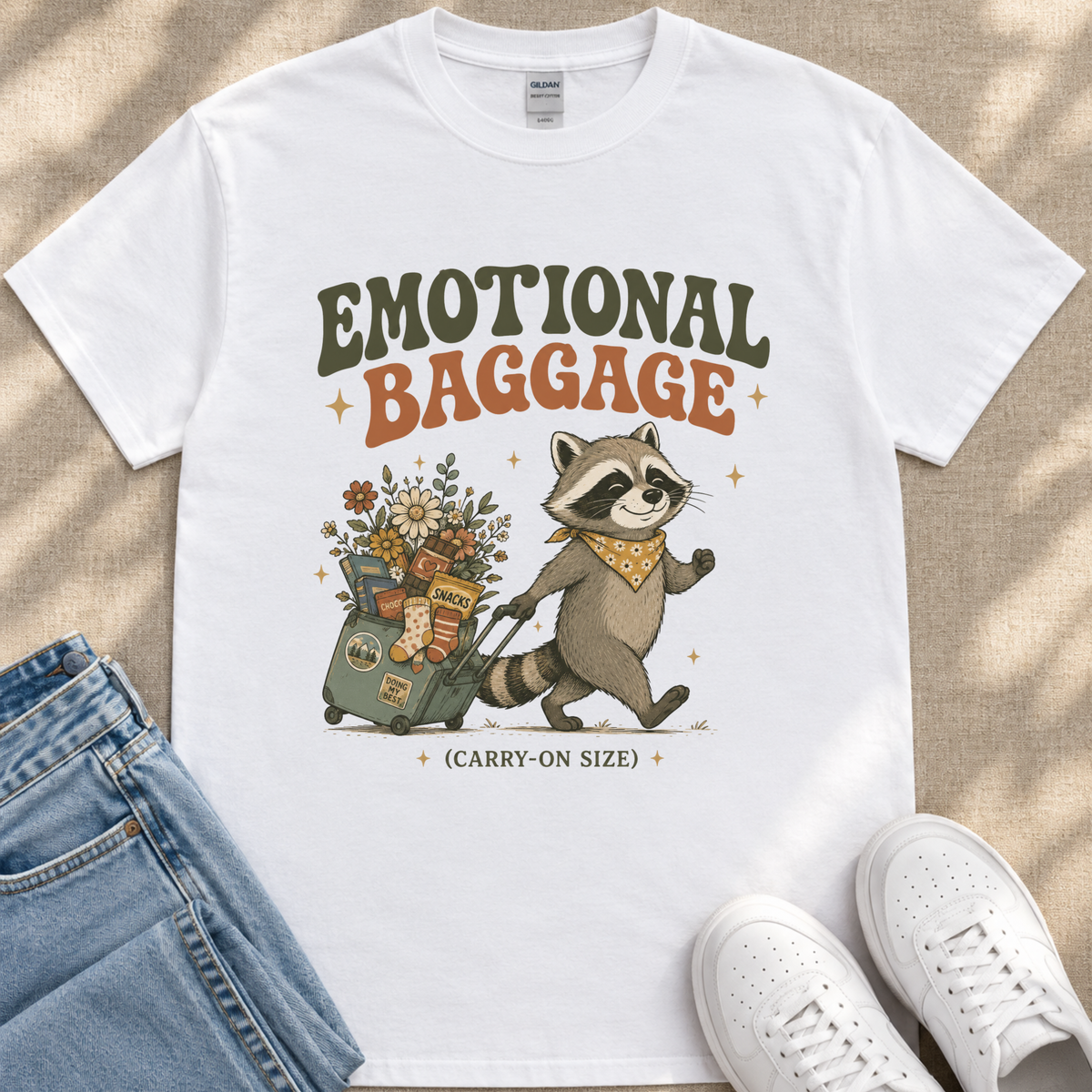 A white t-shirt with a graphic design depicting a raccoon pulling a suitcase filled with various items, accompanied by the text "Emotional Baggage" and "Carry-on Size".