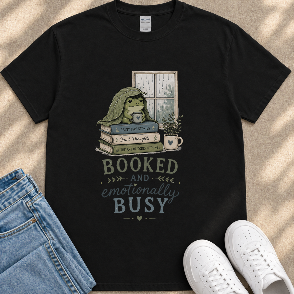 A black t-shirt with a graphic design depicting a frog sitting on a stack of books, with the text "BOOKED AND EMOTIONALLY BUSY" printed on it.