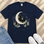 A navy blue t-shirt with a graphic design depicting a black cat sitting on a crescent moon, surrounded by various objects such as a book, a candle, and a plant.