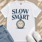 A white t-shirt with the text "SLOW IS SMART" printed on it, along with an illustration of a pocket watch. The t-shirt is placed on a beige surface, and there are a pair of jeans and white sneakers visible in the background.