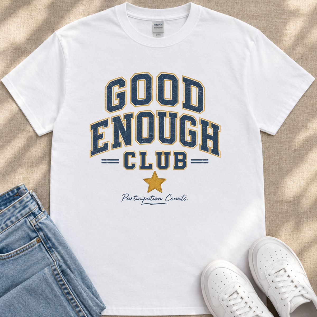 A white t-shirt with the text "GOOD ENOUGH CLUB Participation Counts" printed on it, along with a yellow star symbol. The t-shirt is placed on a beige surface, and there are a pair of blue jeans and white sneakers visible in the image.