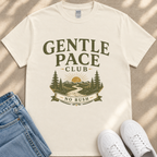 A white t-shirt with a graphic design featuring a mountain landscape and the text "GENTLE PACE CLUB NO RUSH" printed on it. The t-shirt is placed on a beige surface, and there are a pair of blue jeans and white sneakers visible in the image.