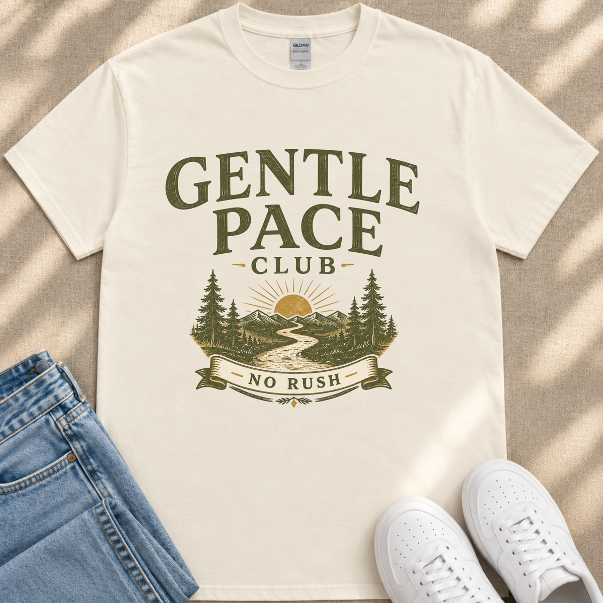A white t-shirt with a graphic design featuring a mountain landscape and the text "GENTLE PACE CLUB NO RUSH" printed on it. The t-shirt is placed on a beige surface, and there are a pair of blue jeans and white sneakers visible in the image.