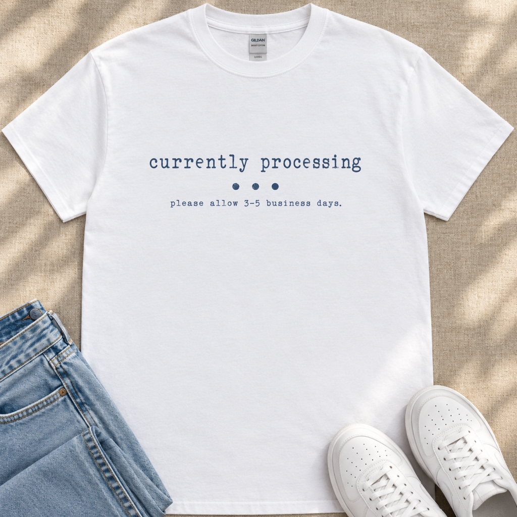A white t-shirt with the text "currently processing" printed on it, along with a message that says "please allow 3-5 business days." The t-shirt is placed on top of a pair of jeans and white sneakers.