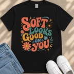 A black t-shirt with a colorful graphic design that says "Soft looks good on you" surrounded by flowers and stars.