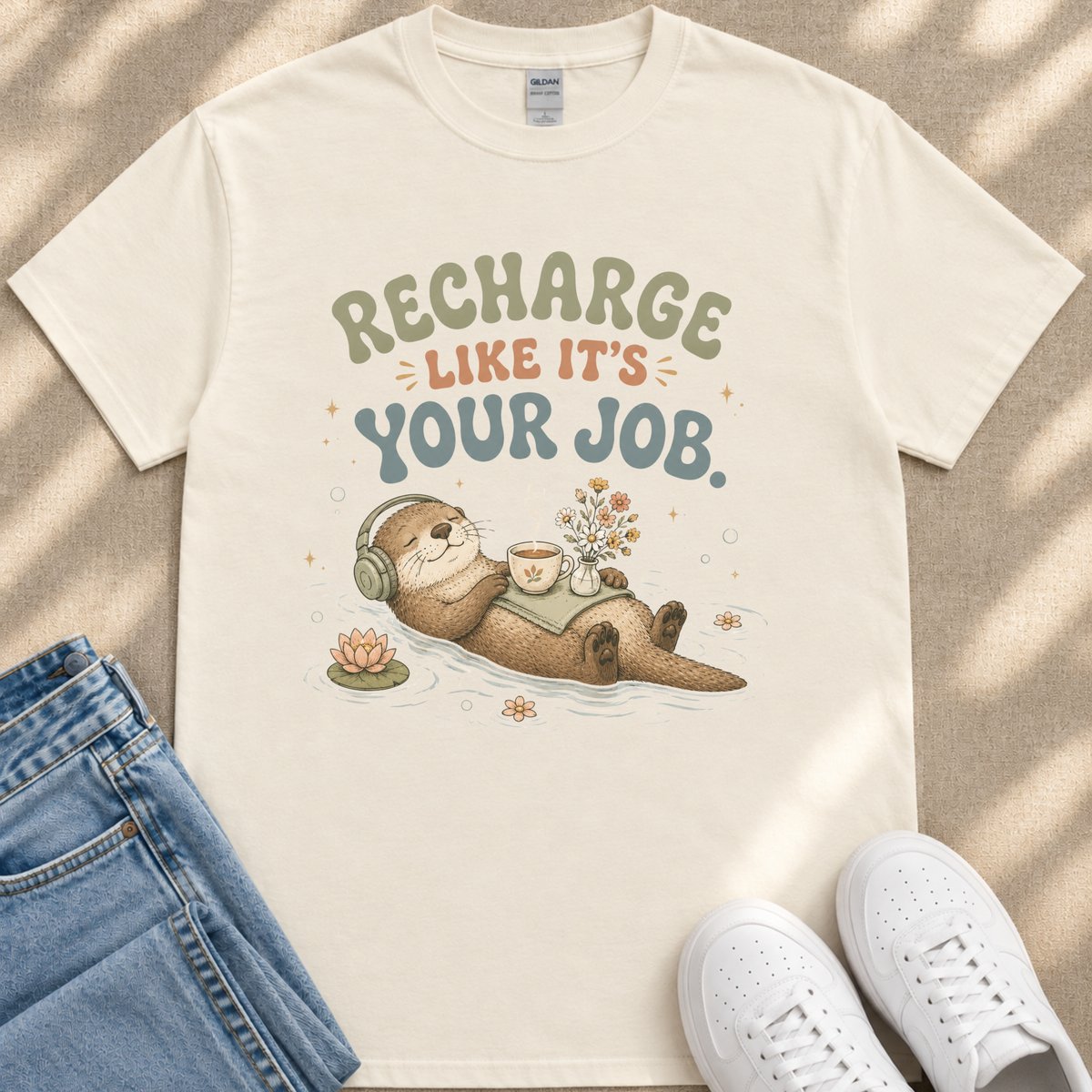 A white t-shirt with a graphic design depicting an otter wearing headphones and holding a cup, surrounded by flowers and a pond, along with the text "Recharge like it's your job."