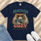 A navy blue t-shirt with a graphic design depicting an opossum wrapped in a blanket, sitting inside a tent with a sign that says "Do not enter". The text "PROTECTED BY COZY ENERGY" is displayed above the opossum.