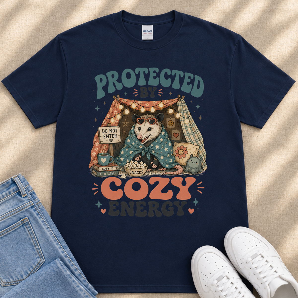 A navy blue t-shirt with a graphic design depicting an opossum wrapped in a blanket, sitting inside a tent with a sign that says "Do not enter". The text "PROTECTED BY COZY ENERGY" is displayed above the opossum.