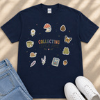 A navy blue t-shirt with a colorful graphic design featuring various objects such as a mushroom, flower, pine cone, butterfly, frog, and other whimsical illustrations, along with the text "Collecting Little Joys".