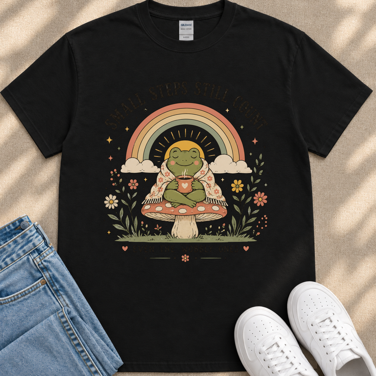 A black t-shirt with a colorful illustration of a frog sitting on a mushroom, holding a cup, surrounded by flowers, leaves, and a rainbow.