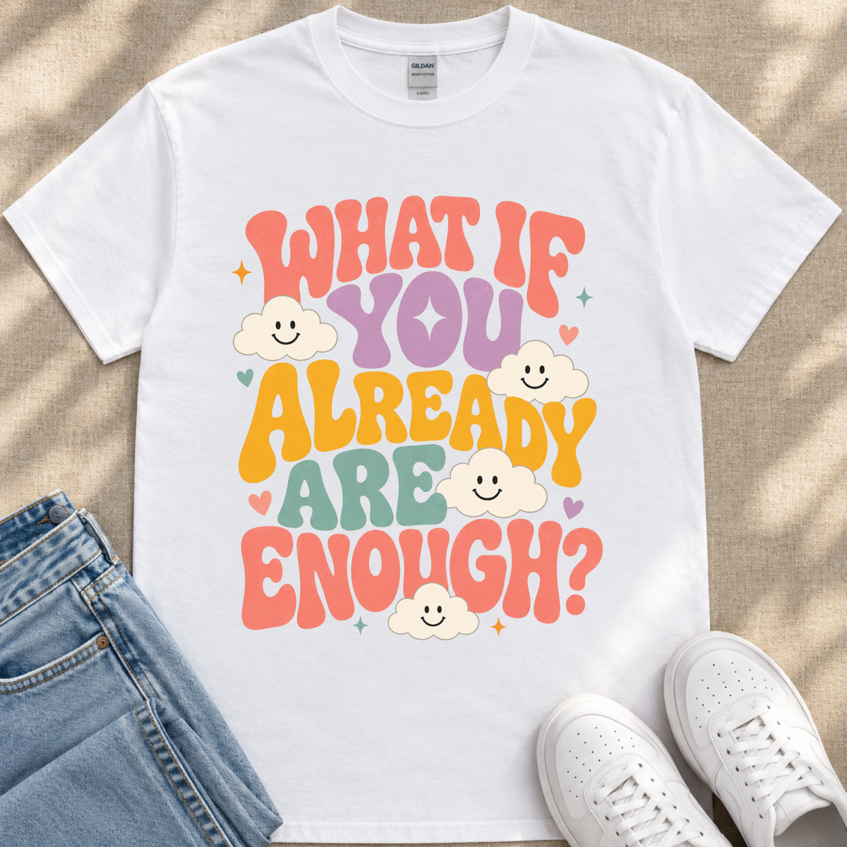 A white t-shirt with a colorful graphic design featuring the text "What if you already are enough?" surrounded by smiling clouds.